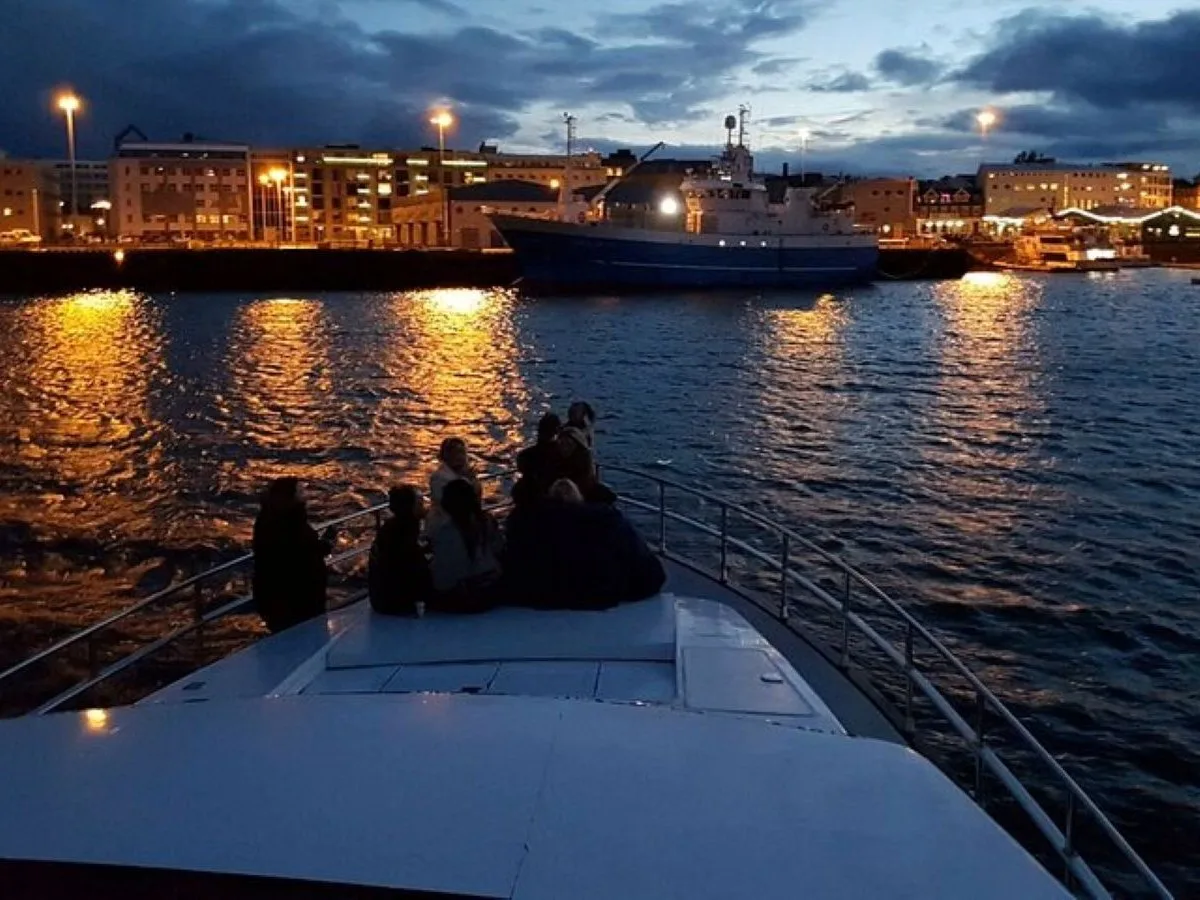 Passengers enjoying the glowing city lights of Reykjavík Old Harbour from the deck of a yacht during an evening cruise at twilight.