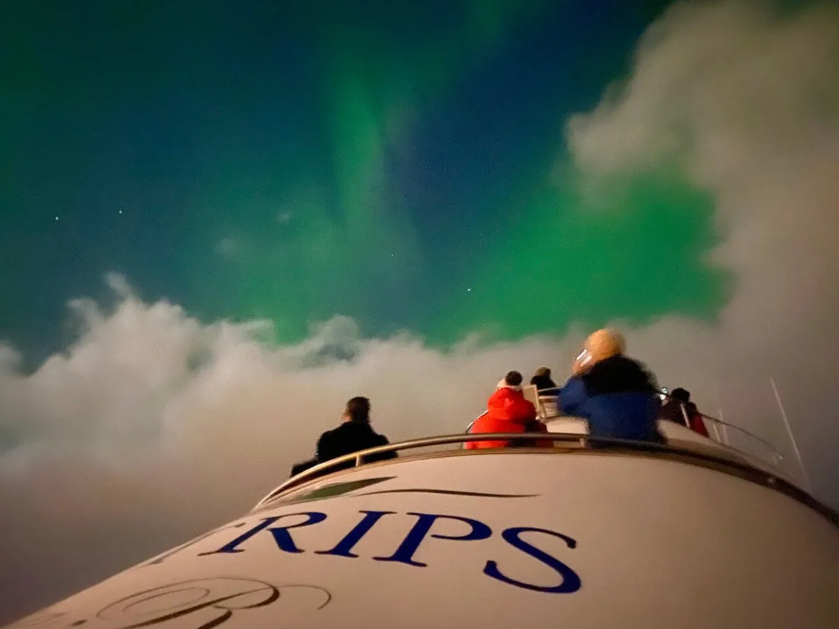Bright green Northern Lights (Aurora Borealis) dancing in the night sky as passengers watch from the observation deck of a yacht cruise in Reykjavík.