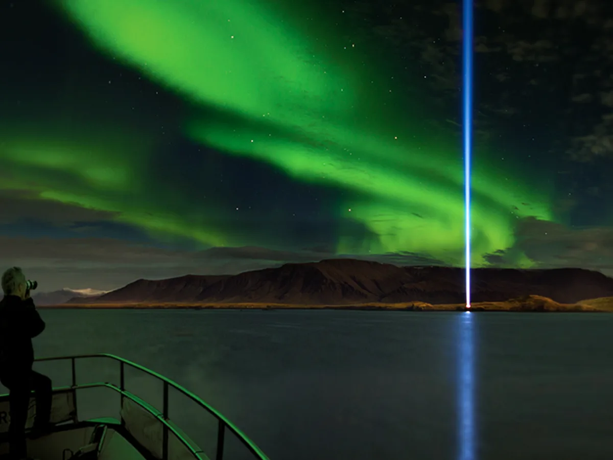 The Imagine Peace Tower blue beam and vibrant Northern Lights seen from the deck of the Amelia Rose yacht near Viðey Island, Reykjavík.